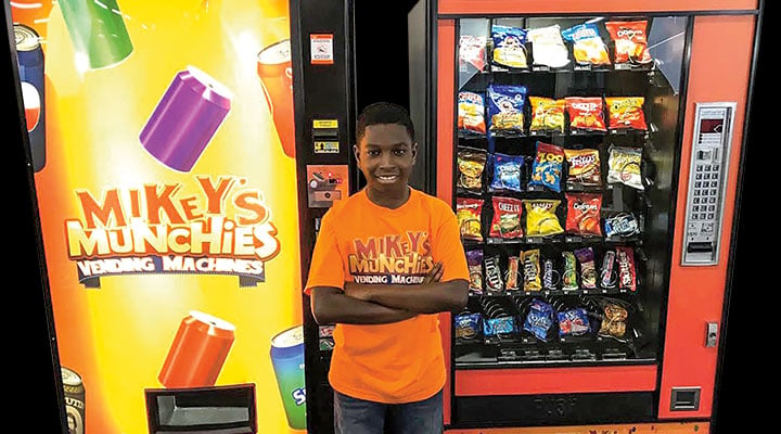 Kid standing in front of two snack vending machines with his business&apos; name on it