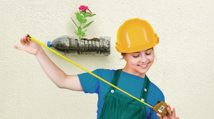 A girl in a yellow hard hat holds a measuring tape and a plastic bottle planter with a flower