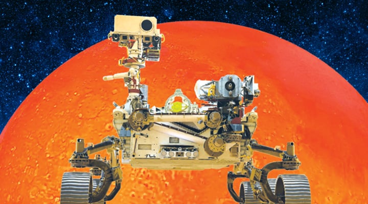 A Mars rover with cameras and instruments sits in front of a large image of Mars